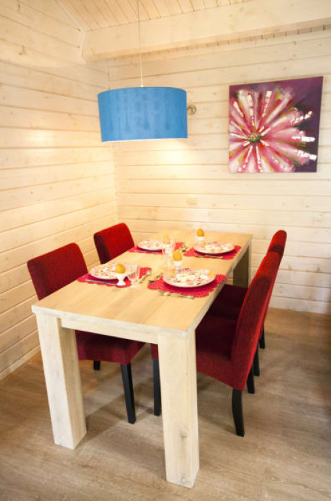 Comedor en una cabaña con paredes de madera, mesa para cuatro, sillas rojas, lámpara azul y cuadro.