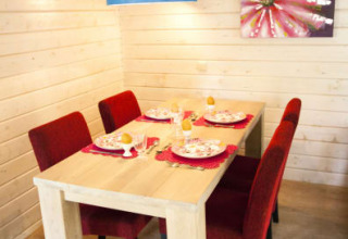 Comedor en una cabaña con paredes de madera, mesa para cuatro, sillas rojas, lámpara azul y cuadro.