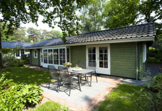 Agradable lodge verde con puertas blancas, área de terraza, mesa y sillas junto a un hermoso jardín boscoso.