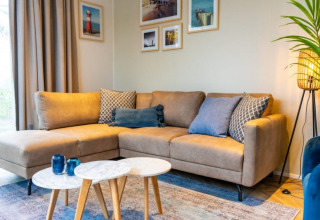 Gezellige woonkamer in Pavilion-lodge met hoekzetel, blauwe kussens, wanddecoratie en staande lamp.