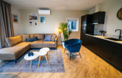Moderne lounge i Pavilion-lodgen med hjørnesofa, blå stol, åbent køkken og dekorative billeder på væggen.