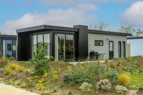 Moderne lodge med store vinduer og terrasse, omgivet af naturlig beplantning under en blå himmel.