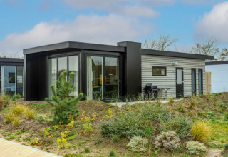 Moderne lodge met grote ramen en terras, omringd door natuurlijke begroeiing en blauwe lucht.