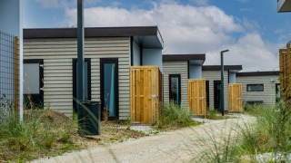 Række af moderne pavillonlodges med træhegn og grussti, omkranset af grønne planter og blå himmel.