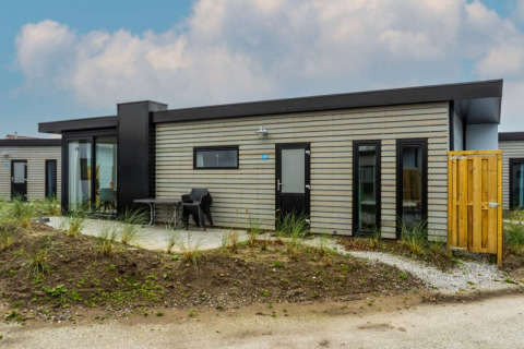 Moderne pavillonlodge med fladt tag, udendørs siddeplads og træbeklædning, omgivet af sand og planter.