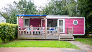 Cabaña Lodge XL rosa con terraza de madera y personas sentadas afuera, rodeadas de árboles verdes.