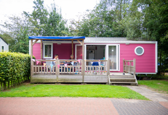 Cabaña Lodge XL rosa con terraza de madera y personas sentadas afuera, rodeadas de árboles verdes.