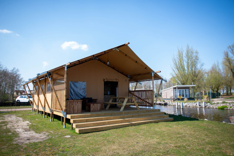 Safari tent lodge with wooden deck and picnic bench by a canal, set in a peaceful natural setting.