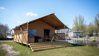 Safari tent lodge with wooden deck and picnic bench by a canal, set in a peaceful natural setting.
