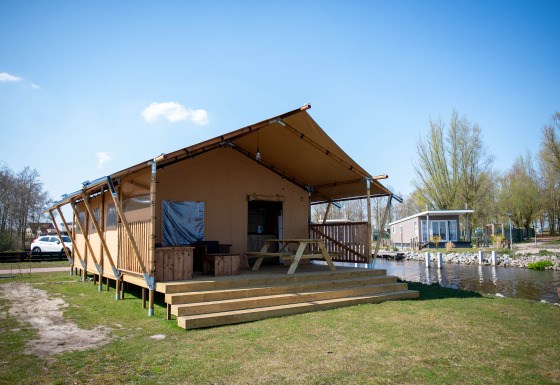 Safari tent lodge with wooden deck and picnic bench by a canal, set in a peaceful natural setting.