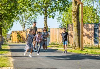 Familie går og leger på en sti i et grønt ferieområde med glamping, omgivet af træer og træhytter.