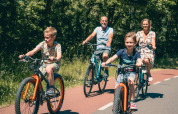 En familie cykler sammen på en solrig dag, omgivet af træer, ved et feriepark med glamping faciliteter.