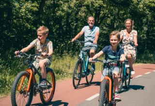 En familie cykler sammen på en solrig dag, omgivet af træer, ved et feriepark med glamping faciliteter.