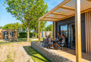 Familie nyder måltid på terrasse ved moderne glampinghytte i solrigt ferieområde med træer og natur.