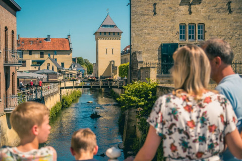 Familia sobre un puente con vistas al río y edificios históricos en un parque de glamping vacacional.