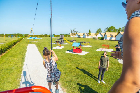 Børn leger på legeplads i en feriepark med glamping, små hytter og solskin i baggrunden.