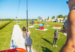 Børn leger på legeplads i en feriepark med glamping, små hytter og solskin i baggrunden.