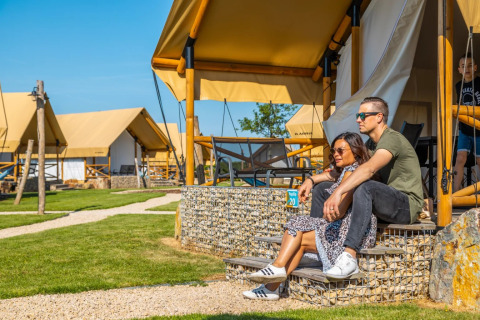 Pareja relajándose fuera de una tienda glamping en un parque vacacional con césped verde y más tiendas.
