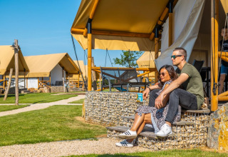 Couple relaxing outside a glamping tent at a holiday park, with green lawns and other tents in view.