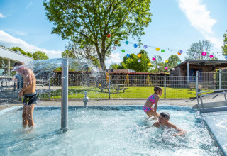 Børn leger i en udendørs swimmingpool med springvand på en solrig dag i et feriepark med glamping.