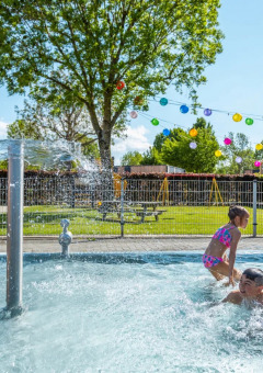 Kinder spielen in einem Außenpool mit Wasserfontäne in einem sonnigen Ferienpark mit Glamping-Angebot.