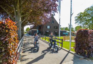 To personer kører el-scootere på en sti i en feriepark med glamping, grønne områder og træer.