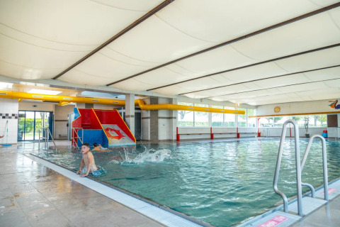 Piscina cubierta en un parque vacacional con tobogán para niños, niños jugando y ventanas panorámicas.