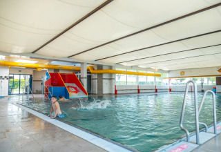 Indendørs swimmingpool i en feriepark med rutschebane, børn der leger, og store vinduer med udsigt udenfor.
