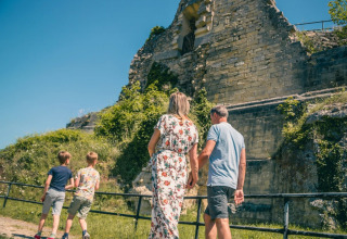 Familie udforsker gamle ruiner nær et glamping-opholdsområde under en klar blå himmel om sommeren.