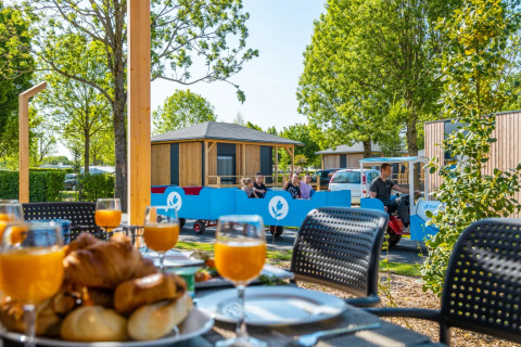 Desayuno con bollería y zumo en la mesa, familia pasea en tren por cabañas glamping en parque vacacional verde.