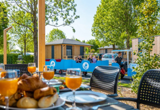 Frühstück mit Gebäck und Saft auf dem Tisch, Familie fährt mit Bummelzug neben Glamping-Hütten im Ferienpark.
