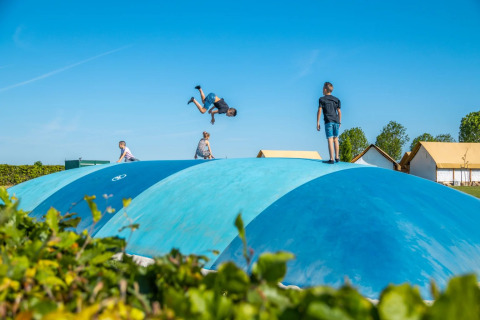Niños juegan en una gran almohadilla azul para saltar en un parque vacacional con tiendas glamping al fondo.