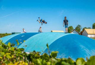 Børn leger på en stor blå hoppepude i en feriepark med glamping-telte i baggrunden under blå himmel.