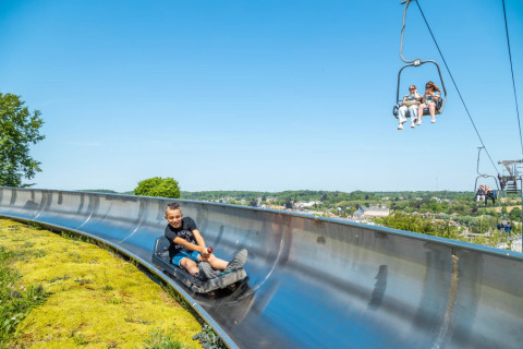 Niño en tobogán de verano y personas en telesilla en parque vacacional de glamping bajo un cielo despejado.