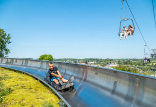 Dreng på sommer rutschebane og mennesker i svævebane i smukke omgivelser på feriepark med glamping.