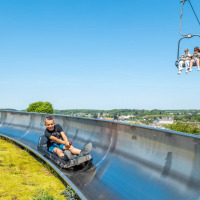 Junge fährt Sommerrodelbahn, während Leute im Sessellift bei einem Glamping-Ferienpark vorbeifahren.