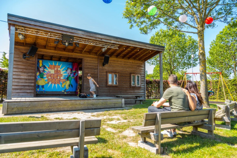 Parc de vacances avec glamping, couple regardant un enfant sur scène extérieure lors d'une journée ensoleillée.