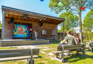 Sceneoptræden på feriepark med glamping, hvor et par ser et barn på scenen i solskinsvejr udenfor.