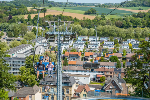 Mensen genieten van een stoeltjeslift boven een vakantiepark met glamping, uitkijkend op huizen en natuur.