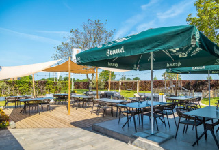 Außenterrasse eines Ferienparks mit Glamping, Tischen unter großen Sonnenschirmen und viel Grün.