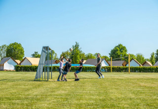 Børn leger fodbold ved siden af glampingtelte i en feriepark på en solrig dag med blå himmel.