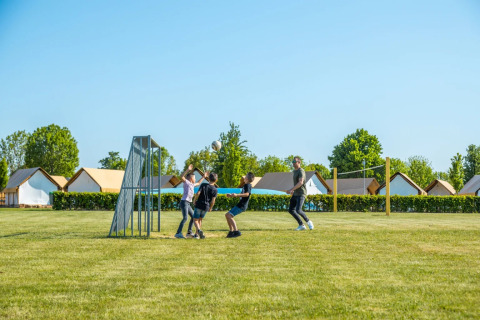 Kinderen spelen voetbal bij glampingtenten op een vakantiepark onder een heldere blauwe lucht.