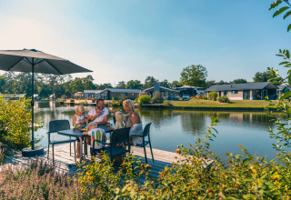 Famille partageant un repas au bord du lac dans un parc de vacances avec hébergements de glamping modernes.