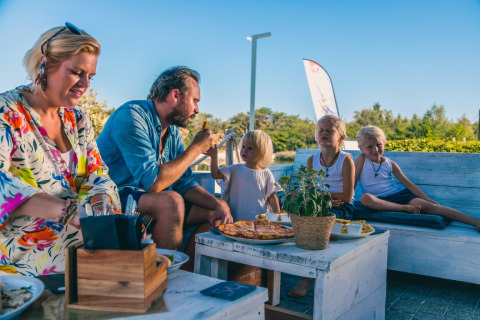 Familie genießt gemeinsam eine Mahlzeit mit Pizza im Freien in einem Glamping-Ferienpark bei Sonnenschein.
