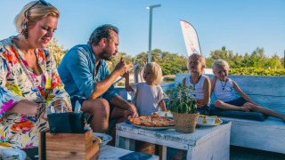 Familie genießt gemeinsam eine Mahlzeit mit Pizza im Freien in einem Glamping-Ferienpark bei Sonnenschein.