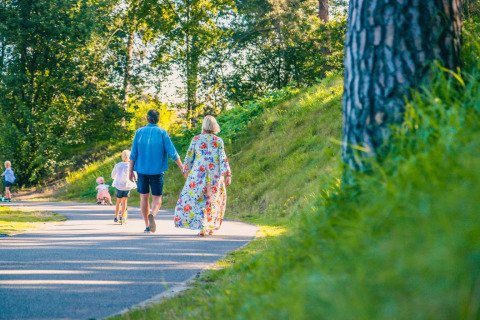 Familie går hånd i hånd på en sti i en feriepark med glamping, omgivet af grønne træer og græs.