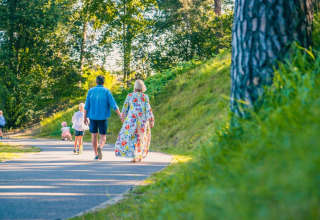 Familie går hånd i hånd på en sti i en feriepark med glamping, omgivet af grønne træer og græs.