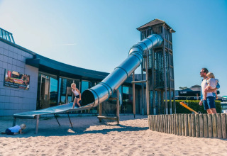 Children play on a large slide in sand at a holiday park offering glamping accommodations on a sunny day.