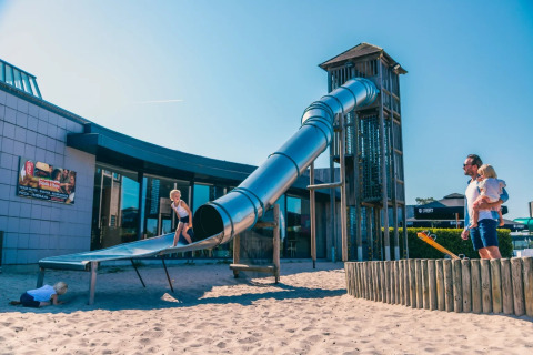 Kinderen spelen op een grote glijbaan met zand bij een vakantiepark met glamping op een zonnige dag.