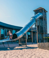 Children play on a large slide in sand at a holiday park offering glamping accommodations on a sunny day.